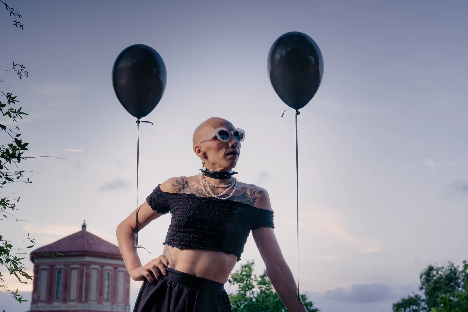 Una persona con el pelo al cero, gafas de sol blancas, un collar de perlas, uno de pinchos y un top negro.